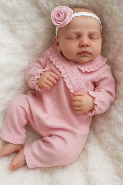 Boneca bebê reborn menina com macacão rosa de babados e faixa de flor na cabeça, deitada em manta branca felpuda com aparência realista e serena.