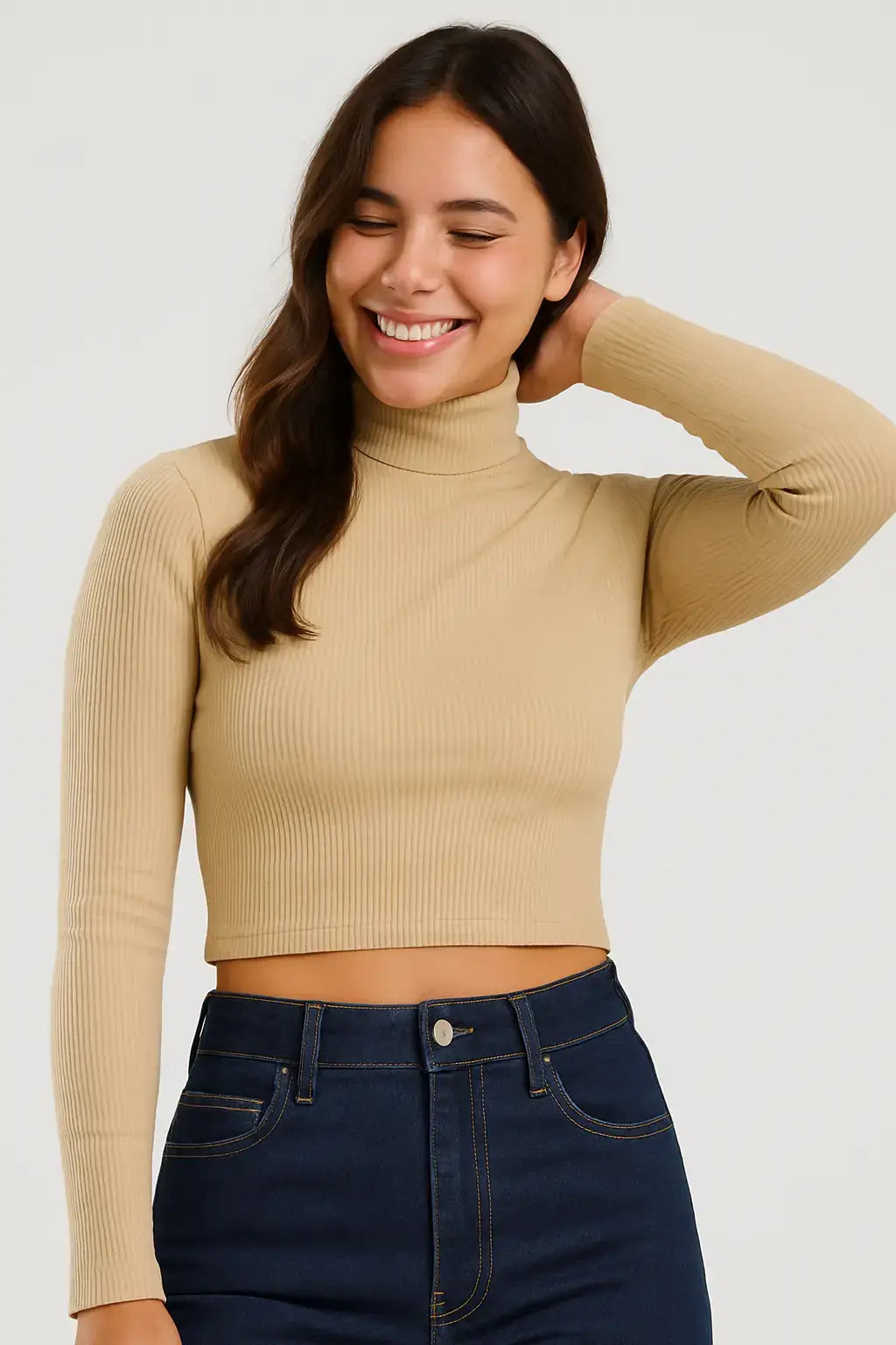 Mulher sorridente com blusa cropped canelada bege de manga longa e gola alta, em pose descontraída com mão no cabelo, usando jeans de cintura alta.