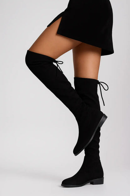 Mulher usando bota preta feminina de cano alto sobre o joelho com amarração traseira, combinada com saia preta curta, ideal para look de inverno elegante.