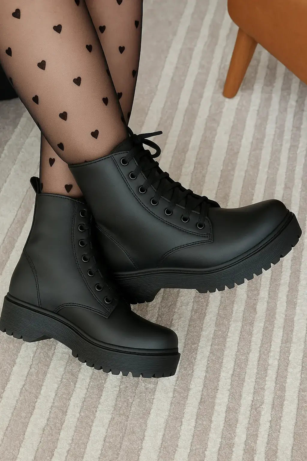 Bota feminina preta de cano curto estilo coturno tratorado com cadarço, usada com meia-calça estampada com corações, sobre tapete bege listrado.