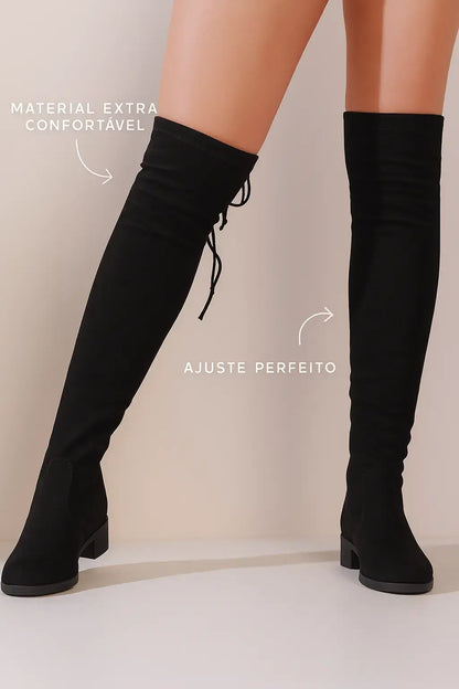 Mulher usando bota over the knee preta com salto baixo e amarração, destacando material confortável e ajuste perfeito sobre fundo bege suave.