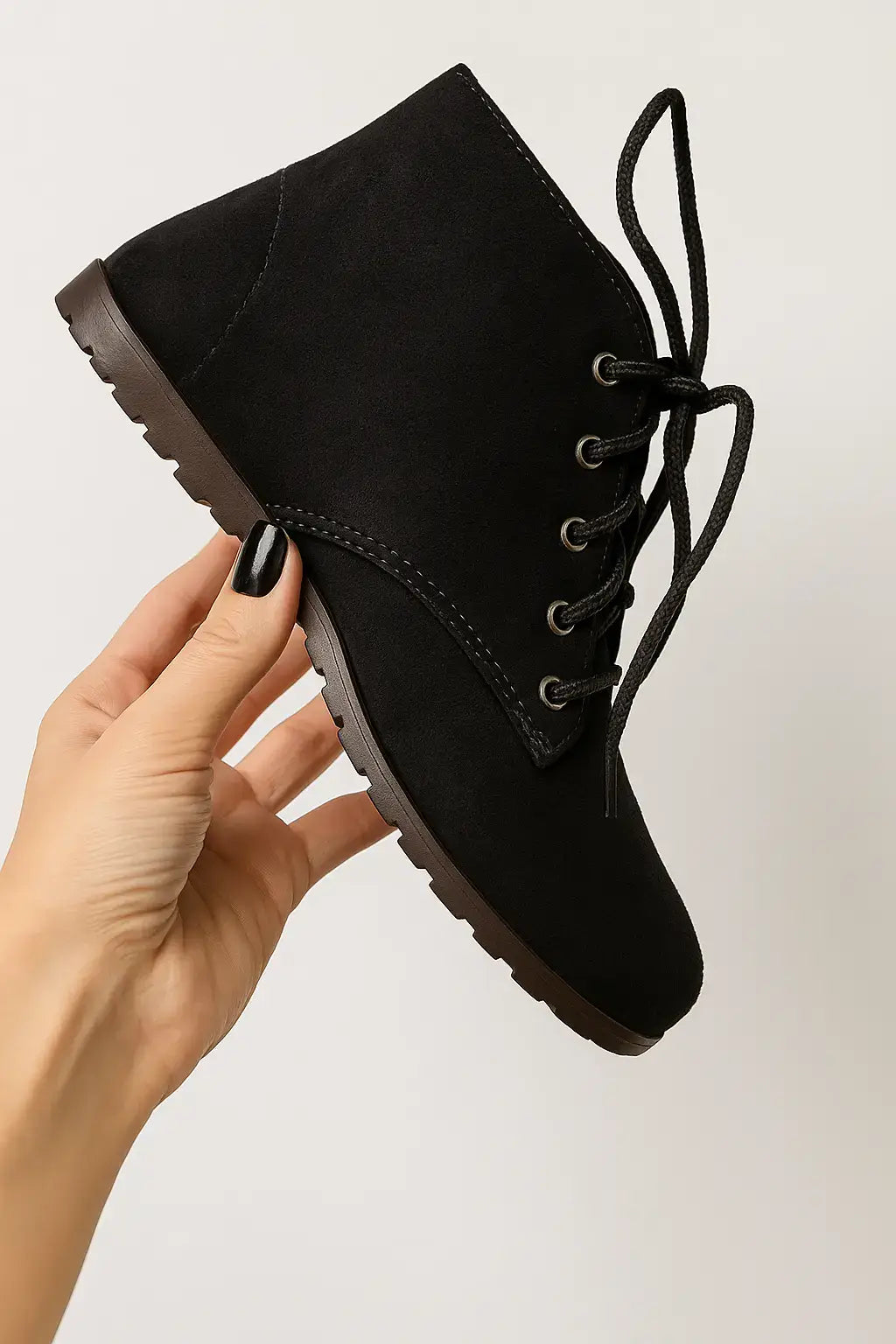 Close da mão segurando bota feminina preta de camurça com cadarço e cano curto, em fundo claro, ideal para vitrines de loja online.