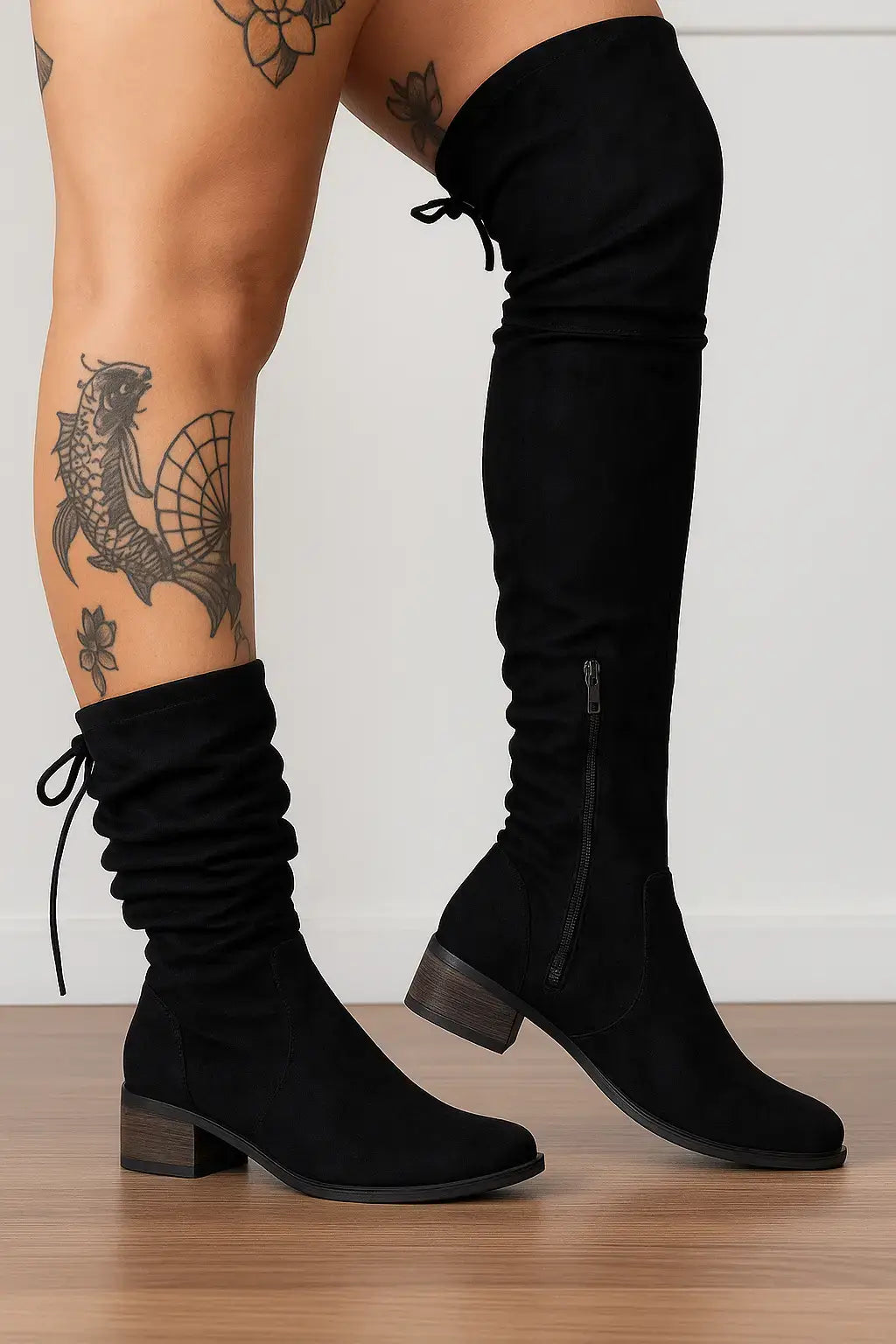 Mulher com tatuagens nas pernas usando bota preta feminina de cano alto com zíper lateral e amarração, sobre fundo neutro e piso de madeira.