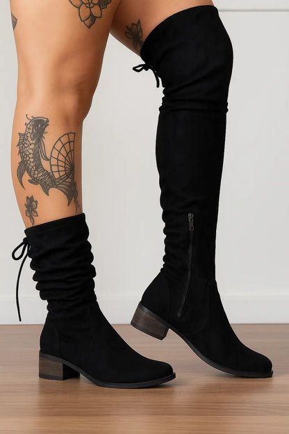 Mulher com tatuagens nas pernas usando bota preta feminina de cano alto com zíper lateral e amarração, sobre fundo neutro e piso de madeira.