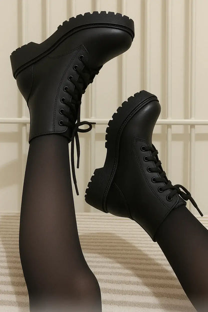 Coturno feminino preto com cano curto e sola tratorada, estilo plataforma, usado com meia-calça preta, em fundo claro com grade metálica vertical.