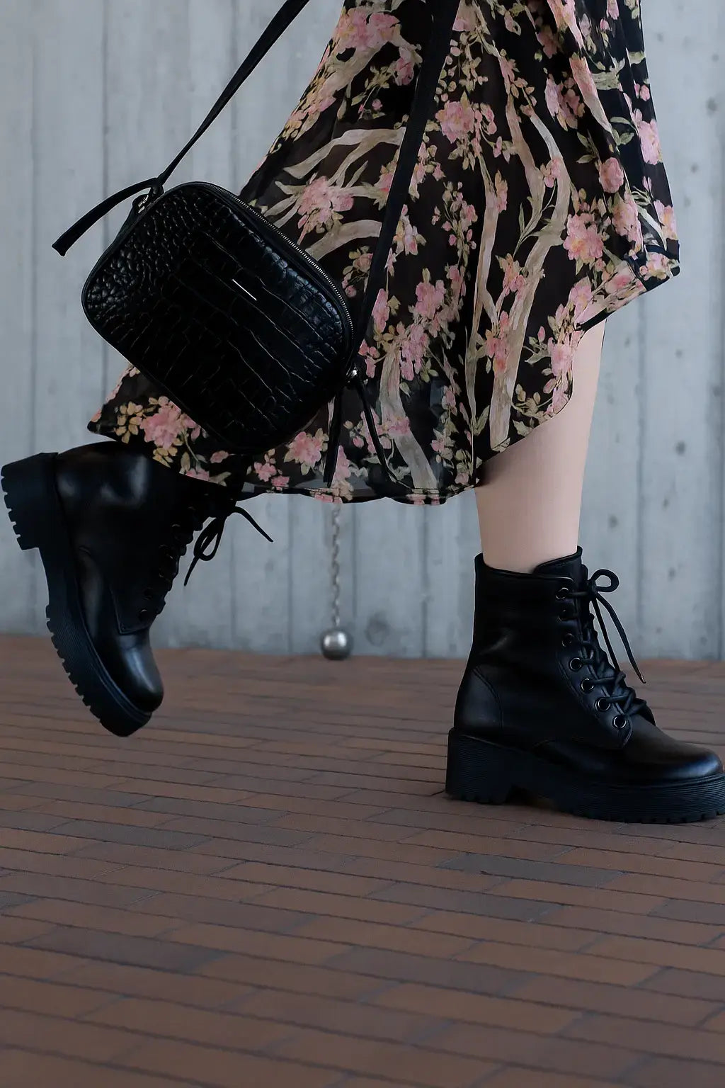 Coturno feminino preto com salto tratorado combinado com vestido floral esvoaçante e bolsa preta com textura de crocodilo, em cenário urbano com parede de concreto e piso de tijolos.