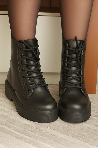 Coturno feminino preto com cano curto, sola alta tratorada e cadarço, usado com meia-calça preta sobre carpete bege listrado, em ambiente interno.