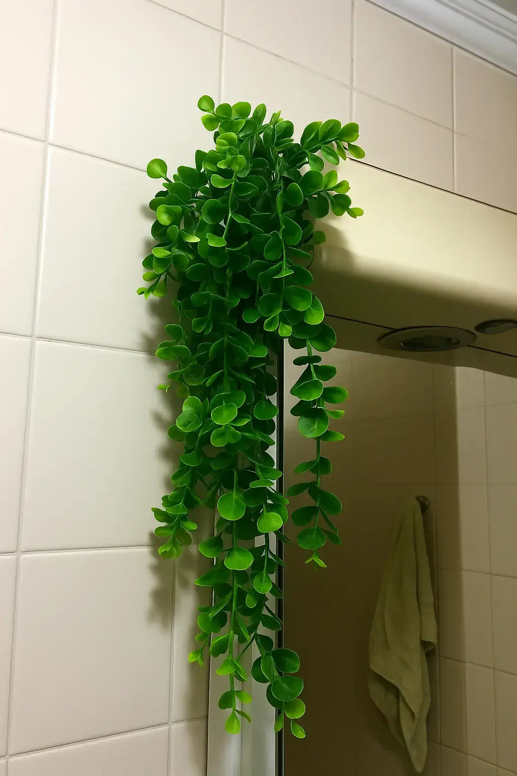 Planta artificial pendente com folhas verdes estilo eucalipto, decorando prateleira de banheiro com revestimento em azulejos bege.