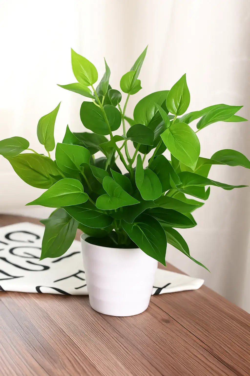 Planta artificial com folhagem verde em vaso branco, sobre mesa de madeira com pano decorativo e cortina ao fundo. Ideal para ambientes internos.
