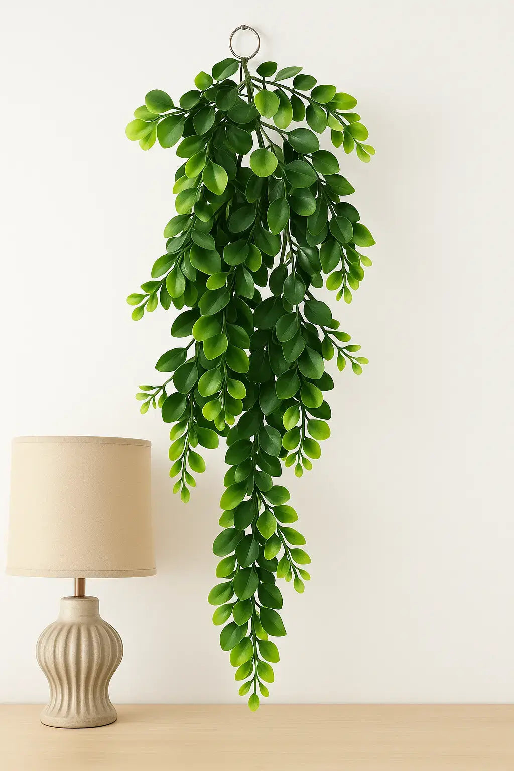 Planta pendente artificial de eucalipto com folhas verdes vibrantes, fixada na parede acima de um abajur bege, ideal para decoração moderna de interiores.